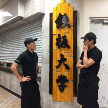 「鉄板大学」共同経営者の牧野弘宗さん（左）と矢澤佑冴さん（右）。木の看板は、牧野さん達が手作りしたもの